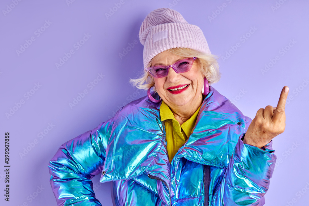 senior lady shows the middle finger and smiles, wearing bright coat and hat, in stylish wear isolated over purple background senior lady shows the middle finger and smiles, wearing bright coat and hat, in stylish wear isolated over purple background