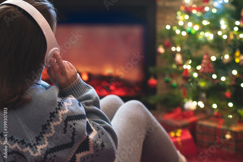 Alone sad woman in headphones sitting and warming at winter evening near fireplace flame and  christmas tree.