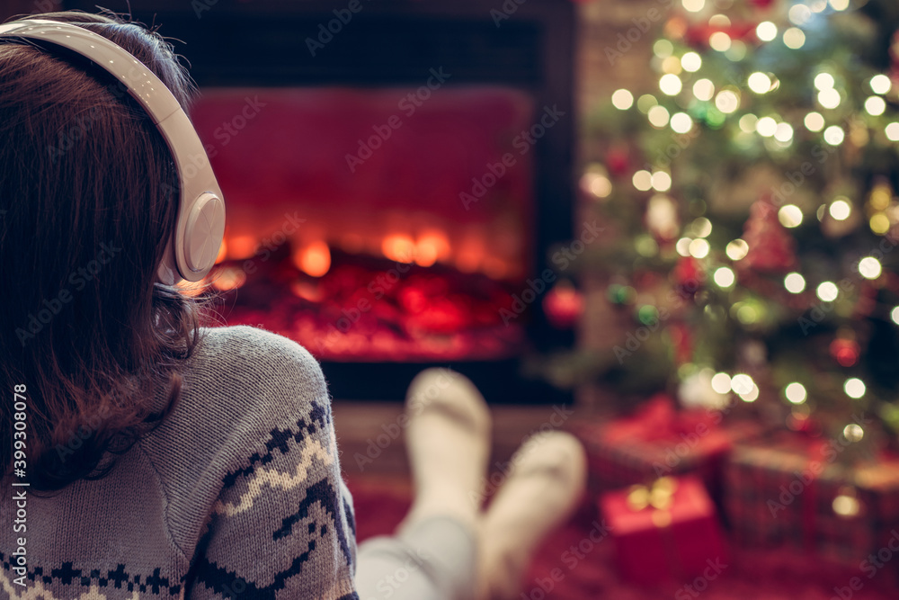 Woman in headphones sitting and warming at winter evening near fireplace flame and christmas tree. Woman in headphones sitting and warming at winter evening near fireplace flame and christmas tree.