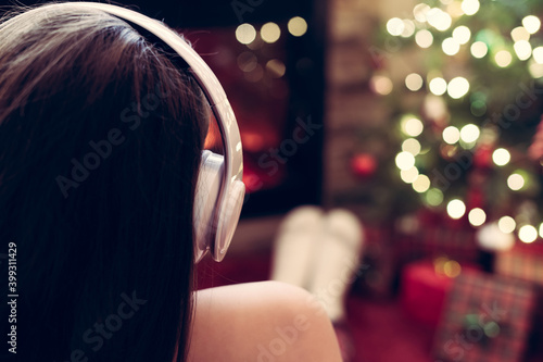 Alone sad woman in headphones sitting and warming at winter evening near fireplace flame and christmas tree.
