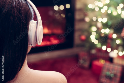 Woman in headphones sitting and warming at winter evening near fireplace flame and christmas tree.
