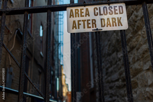 Area Closed After Dark sign on metal plate is attached to the metal railings of a door in front of a vintage passageway at an urban location. A narrow alley is seen in blurred background behind fence.