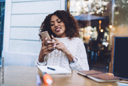 Smiling dark skinned curly hipster girl enjoying 4G connection for watching video on smartphone outdoors, positive african american millennial woman making book on web pages via mobile phone