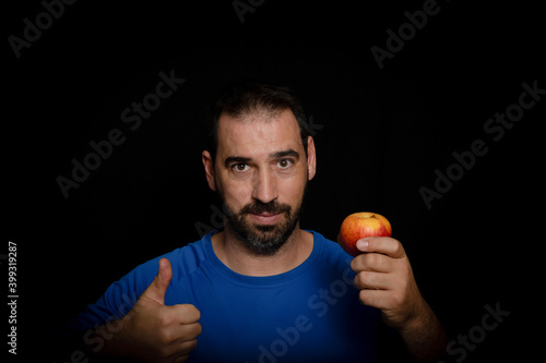 Bearded man dressed in blue t-shirt and holding an apple posing on black background
