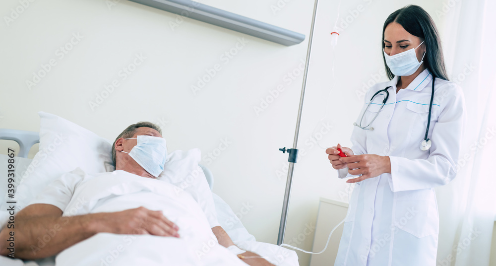 A confident doctor woman in a safety medical mask is examining a mature patient in the mask while he lying in the ward. A confident doctor woman in a safety medical mask is examining a mature patient in the mask while he lying in the ward.