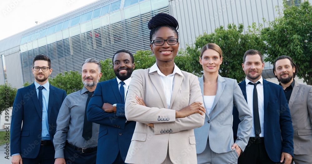 Portrait of successful African American female young boss with team of males and females company workers. Mixed-races businessmen and businesswomen smiling to camera outdoors at business center. Portrait of successful African American female young boss with team of males and females company workers. Mixed-races businessmen and businesswomen smiling to camera outdoors at business center.
