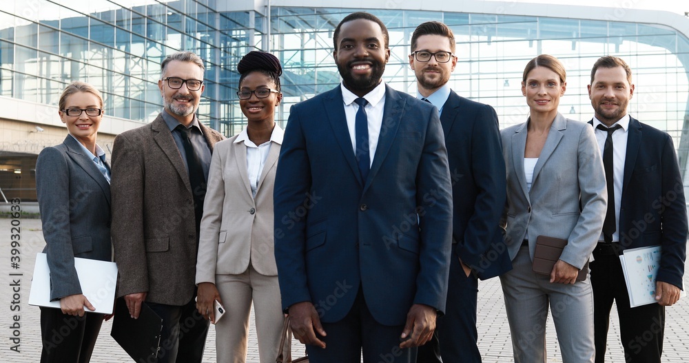 Portrait of successful African American male young boss with his team of males and females company workers. Mixed-races businessmen and businesswomen smiling to camera outdoor at business center.