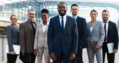 Portrait of successful African American male young boss with his team of males and females company workers. Mixed-races businessmen and businesswomen smiling to camera outdoor at business center.