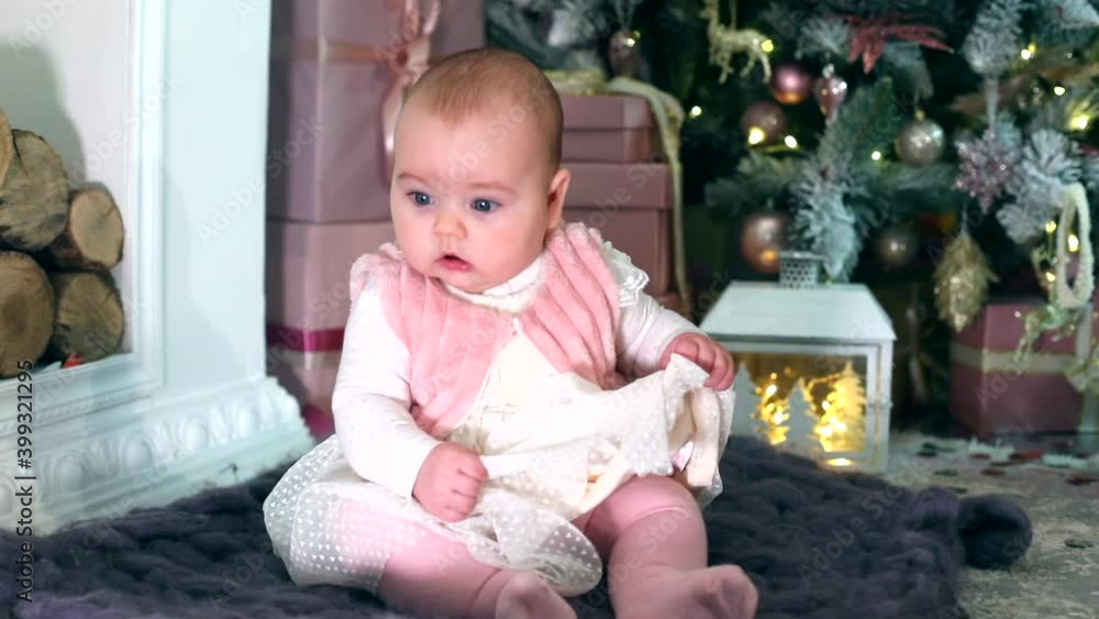 Excited little baby girl in princess dress sitting on carpet near Christmas tree enjoying New Year celebration at home with family. Caucasian female child. Home modern interior. Lights on background Excited little baby girl in princess dress sitting on carpet near Christmas tree enjoying New Year celebration at home with family. Caucasian female child. Home modern interior. Lights on background