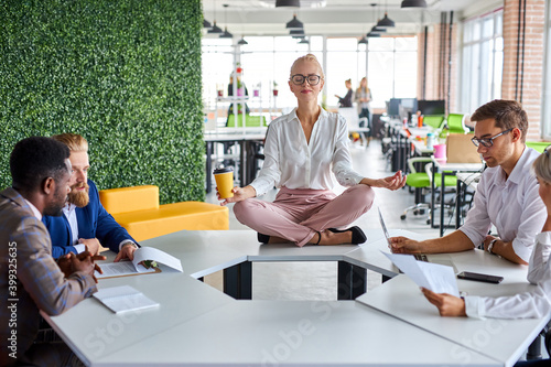 young caucasian woman meditating in office while colleagues are working, keep calm, meditate. sits on table with crossed legs