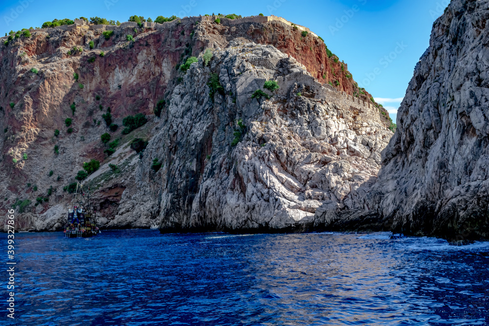 High rock with a steep stone cliff in the Mediterranean Sea with the ancient Alanya Castle at the top (Turkey). Seascape with vintage pirate ship at the foot of the mountain in blue water