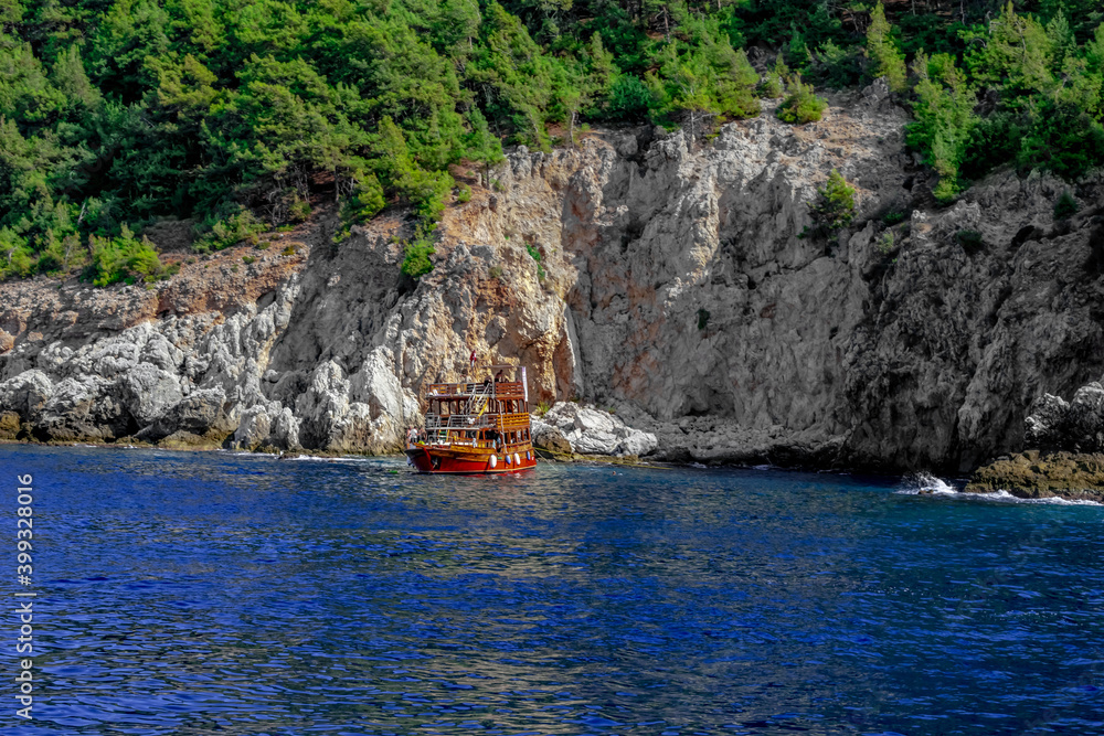Red and orange wooden boat in the blue water of the Mediterranean Sea against the background of a steep cliff and forest on a mountain in Alanya (Turkey). Boat or yacht cruises in a Turkish resort