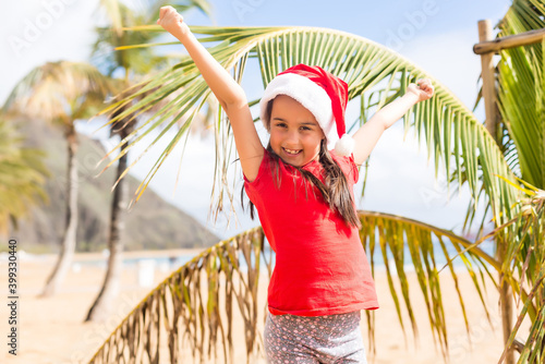 Little girl in santa hat standing next to palm tree on beach