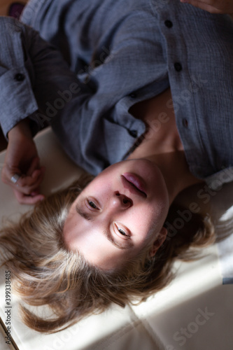 Close-up portrait from top of beautiful young blonde woman without make-up in gray linen shirt is lying on sofa in interior with sunlight