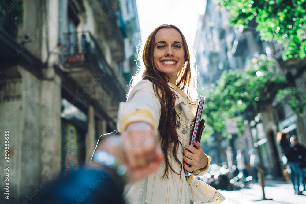 Portrait of cheerful hipster girl rejoicing at urban setting in city during summer vacations or honeymoon, happy Caucasian female tourist with red hair holding hand for follow and laughing at camera