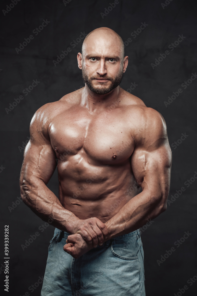 Muscular and bald caucasian man with perfect abs and huge biceps poses in dark background looking at camera with serious face.