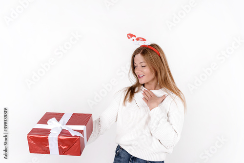 Beautiful young woman wearing white sweater, holding christmas gift box in excitement, wearing funny head hoop, isolated on white background