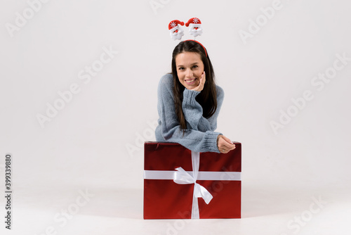 Beautiful young woman wearing blue sweater, sitting near big christmas gift box, wearing funny head hoop, isolated on white background