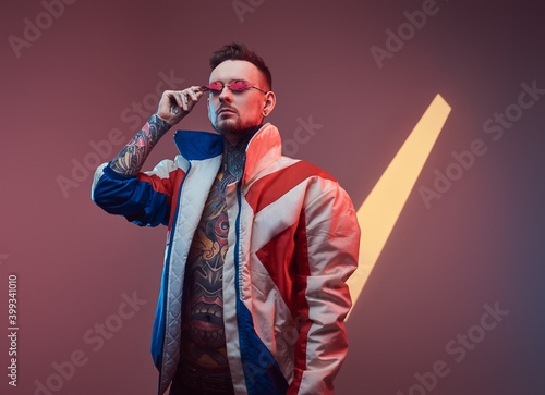 Fashionable and bearded hipster person with sunglasses and tattooed nude body dressed in warm winter coat poses in colourful background with serious face. photo