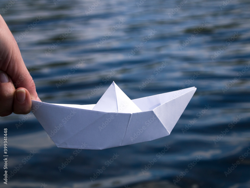 A hand is holding a white paper boat above the water. Origami ship - symbol of new beginnings. The concept of starting a new project or achieve a new goal. The beginning of the way.