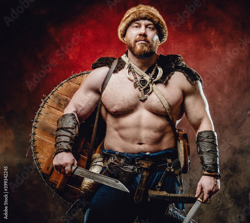 Armed with two knives muscular and shirtless northern barbarian with hat and shield on his back poses in dark red background.