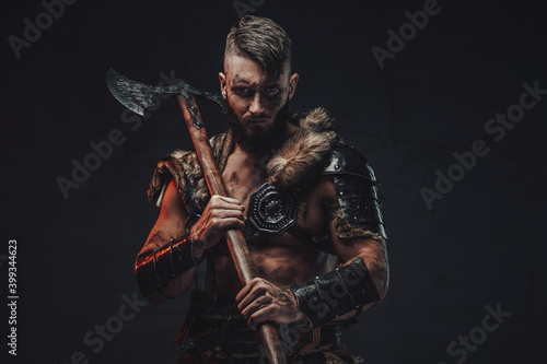 Dressed in light armour with fur scandinavian seafarer armed with two handed axe poses in dark background.