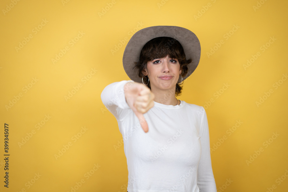 Young caucasian woman wearing hat over isolated yellow background with angry face, negative sign showing dislike with thumb down Young caucasian woman wearing hat over isolated yellow background with angry face, negative sign showing dislike with thumb down