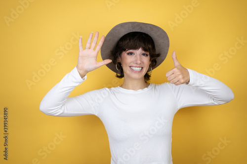 Young caucasian woman wearing hat over isolated yellow background showing and pointing up with fingers number six while smiling confident and happy