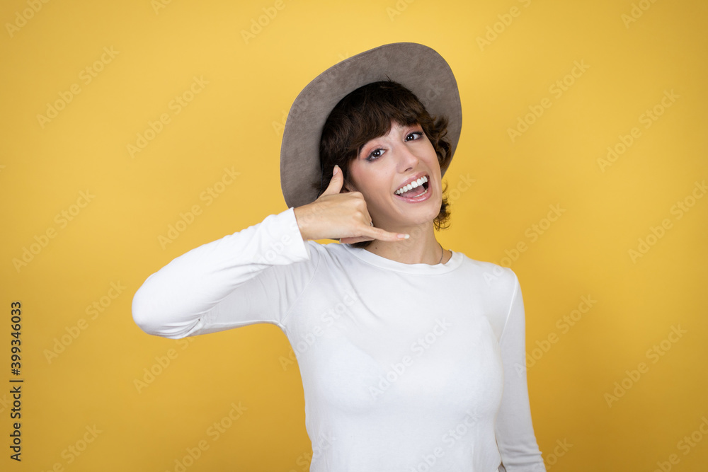 Young caucasian woman wearing hat over isolated yellow background smiling doing phone gesture with hand and fingers like talking on the telephone Young caucasian woman wearing hat over isolated yellow background smiling doing phone gesture with hand and fingers like talking on the telephone