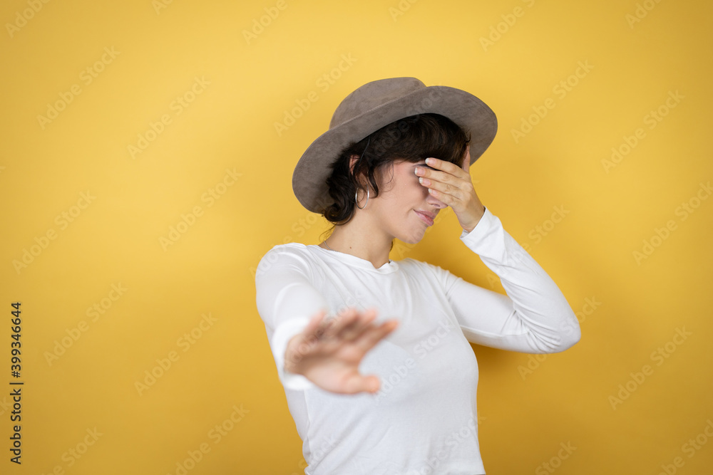 Young caucasian woman wearing hat over isolated yellow background covering eyes with hands and doing stop gesture with sad and fear expression. Embarrassed and negative concept. Young caucasian woman wearing hat over isolated yellow background covering eyes with hands and doing stop gesture with sad and fear expression. Embarrassed and negative concept.
