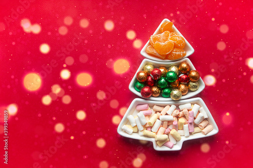 on a red background there is a tray in the form of a Christmas tree. sweets in the tray: marshmallows, round candies in foil and tangerine slices