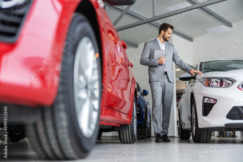 Car seller walking around car salon and holding tablet. There are many new cars ready to be sold.