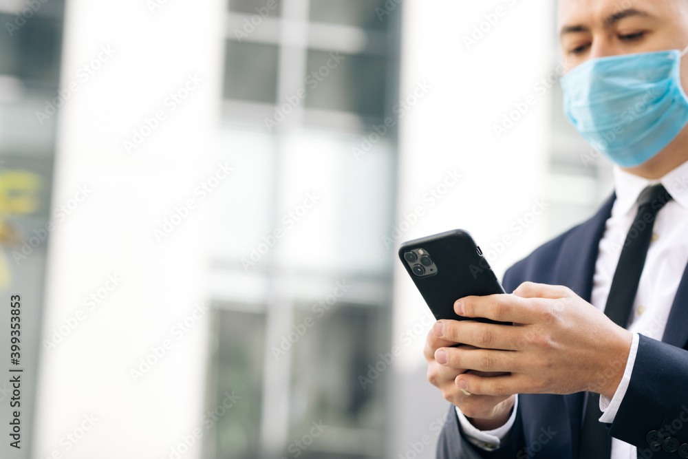 Man wearing protective face mask in the street looking at smartphone surfing social media app. Young man ordering taxi in mobile application, web surfing typing message