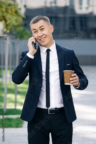 European businessman talking on mobile phone walking near office building background. Man is in dialogue, smiling, holding coffee cup on summer day. © uflypro