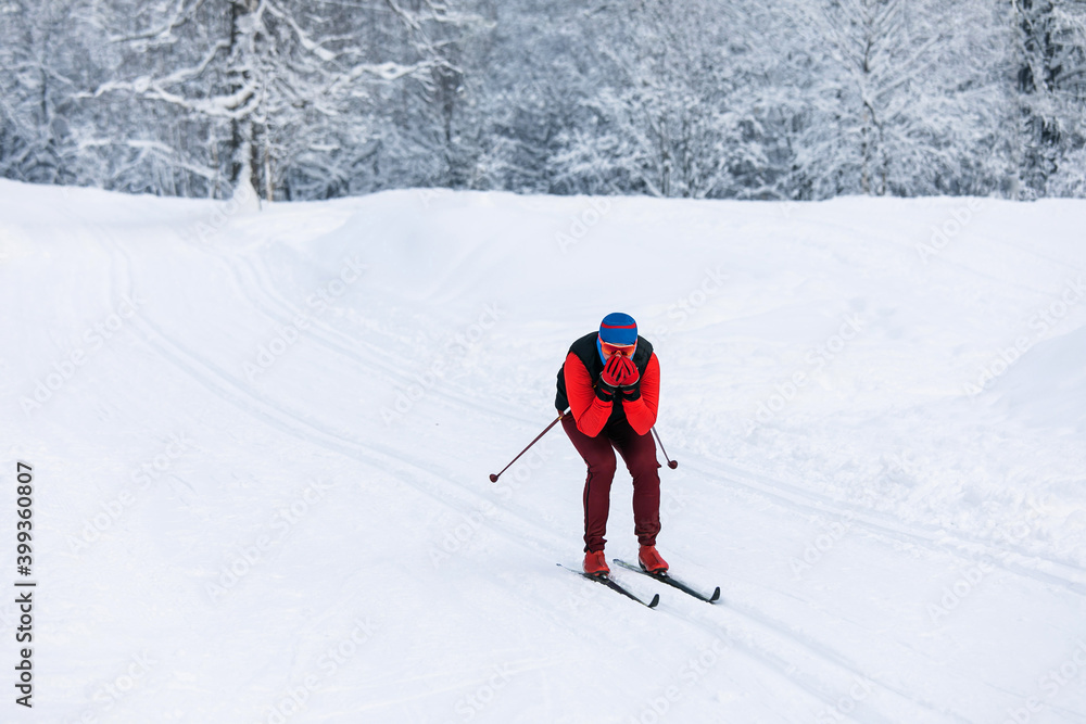 A skier in a red tracksuit, blue balaclava, and safety glasses riding fast downhill, covering his face with his hands from the cold wind with a snow-covered forest background on a cloudy day A skier in a red tracksuit, blue balaclava, and safety glasses riding fast downhill, covering his face with his hands from the cold wind with a snow-covered forest background on a cloudy day