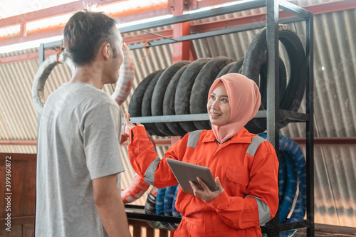 veiled Asian female mechanic uses a digital tablet while chatting with male consumers standing in the background of a tire rack