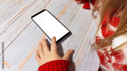 The child is sitting at a table with a phone with a white screen. A girl with a phone layout. In new year's clothing. New Year