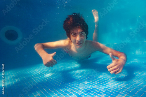 Happy handsome boy float underwater with smiling portrait in the pool show thumb up hand gesture