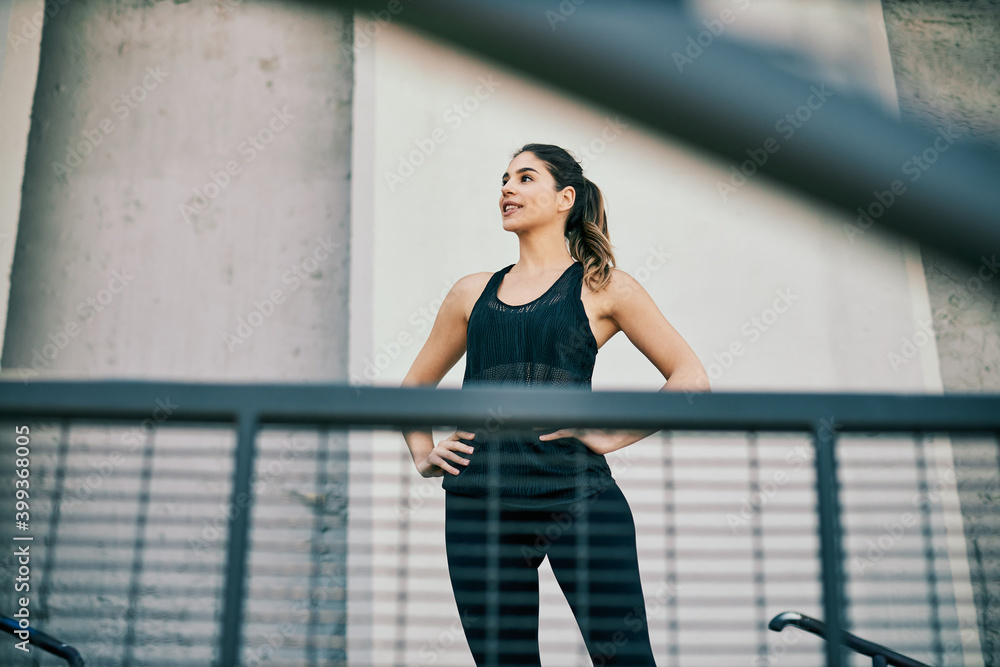 Fototapeta premium Young attractive female runner in shape standing with hands on the hips and taking a break after running in urban exterior.