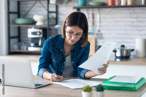 Pretty young business woman working with computer while consulting some invoices and documents in the kitchen at home. photo