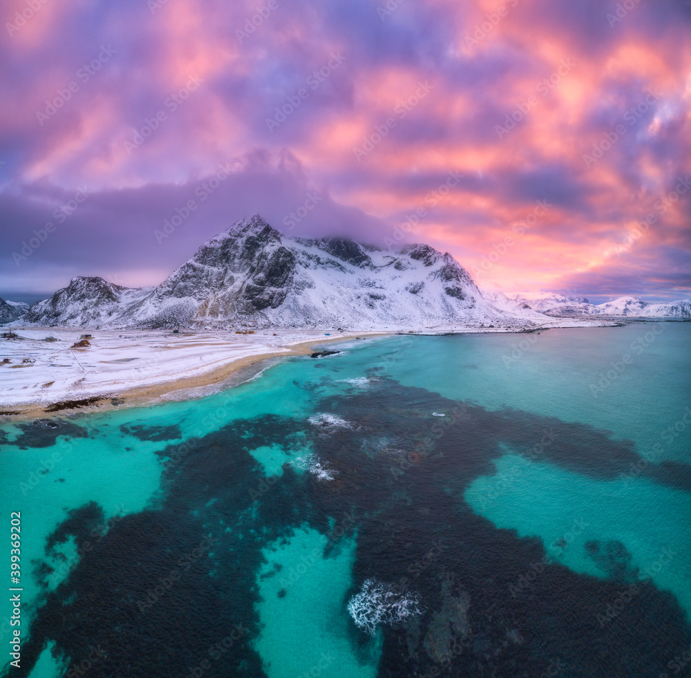 Nordic sandy beach with blue sea in winter at sunset in Lofoten islands, Norway. Landscape with snowy mountains, dramatic sky with orange clouds, water, village with buildings, walking people. Nature
