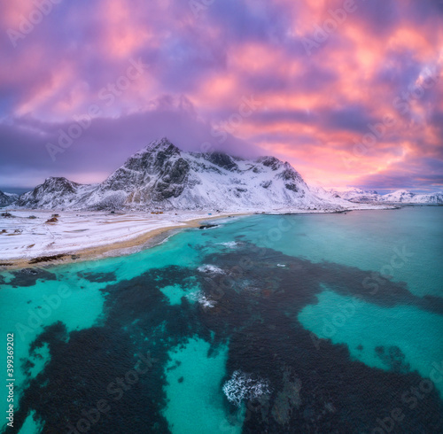 Nordic sandy beach with blue sea in winter at sunset in Lofoten islands, Norway. Landscape with snowy mountains, dramatic sky with orange clouds, water, village with buildings, walking people. Nature