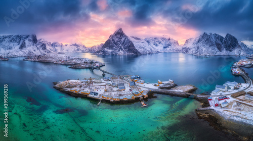 Aerial view of blue sea, snowy mountains, rocks, village, buildings, rorbu, road, bridge, colorful cloudy sky at sunset in winter. Hamnoy in Lofoten islands, Norway. Panoramic landscape. Top view