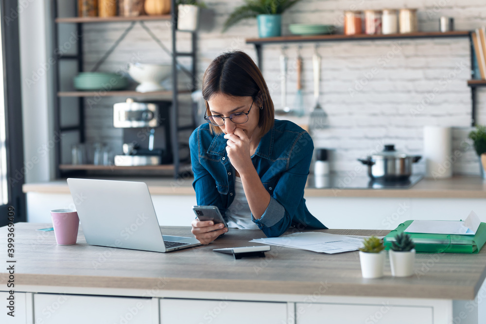 Concentrated young business woman using her mobile phone while working with computer in the kitchen at home. Concentrated young business woman using her mobile phone while working with computer in the kitchen at home.