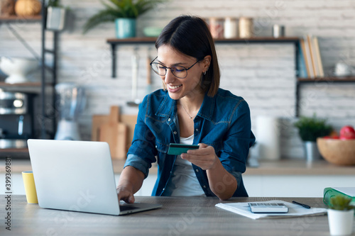 Cute young woman holding white credit card for shopping online with computer while sitting in the kitchen at home. photo