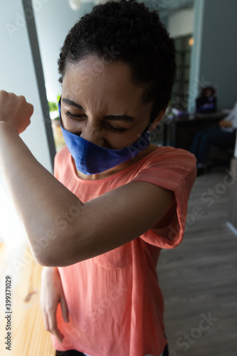 Mixed race woman wearing face mask sneezing into her elbow