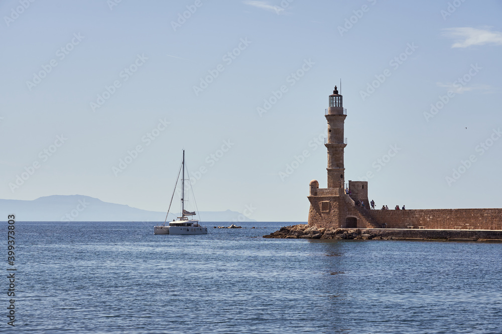 An ancient lighthouse in the port of Hania in Crete and a yacht with a clear and blue sky in the background.