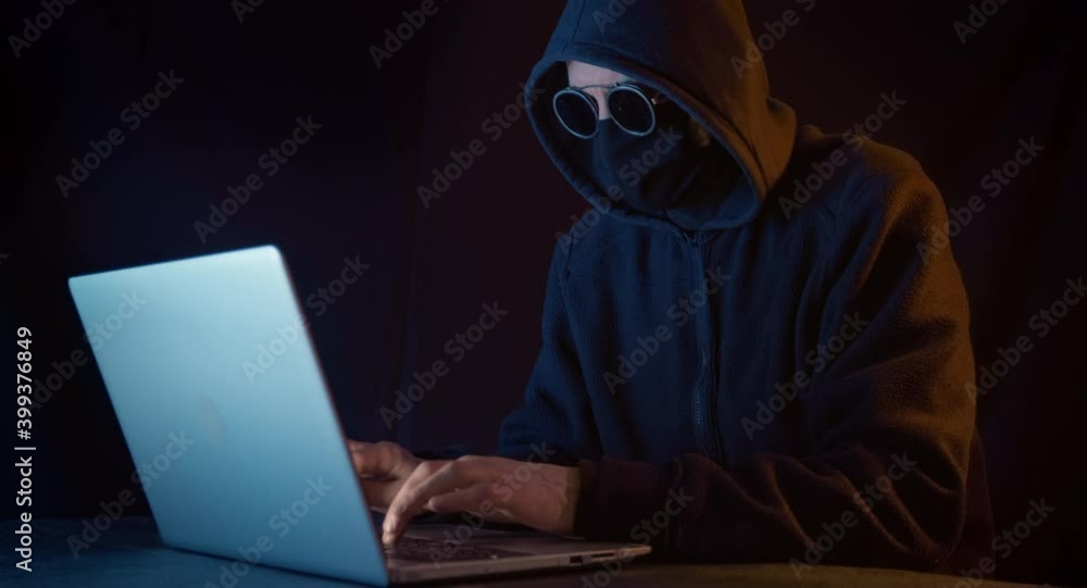 A hacker with a closed face sits at a laptop in a dark room. A hacker with a closed face sits at a laptop in a dark room.