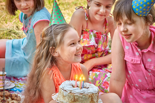 Beautiful happy little girl holding a cake with burning candles in the company of her friends. Celebrate your birthday in nature.