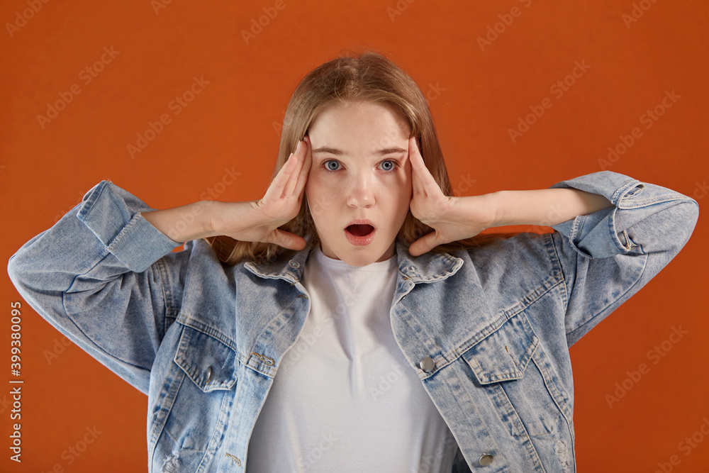 A terrified girl looks at the camera and holds her head in her hands. Bright orange background. A terrified girl looks at the camera and holds her head in her hands. Bright orange background.